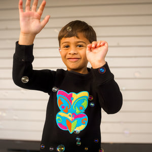 Child wearing a black sweater with a colorful taniwha design, waving in front of a metallic background.